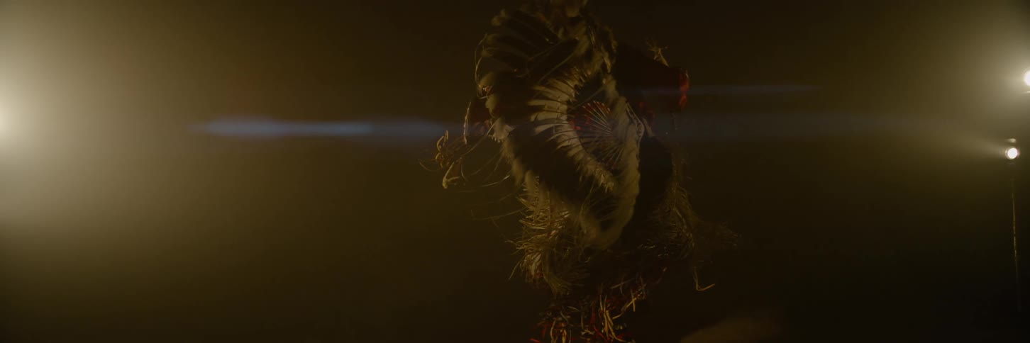 An elderly man dances in elaborate native american ceremonial attire adorned with large feathers and intricate details in a dark hazy environment with bright lights on either side