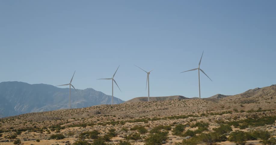 Wind turbines stand on a barren scrubcovered hillside with mountains in the background under a clear blue sky