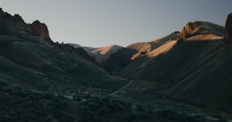 Golden sunlight illuminates rugged mountain peaks and casts long shadows across a winding dirt road in a deep valley