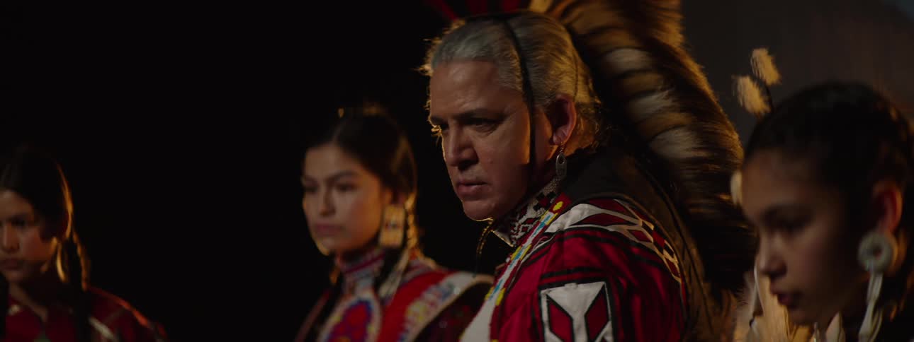 An american native tribe group including a man and several young ladies all dressed in traditional native attire showcasing their culture on a stage