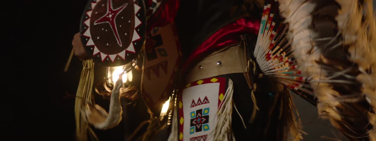 A closeup of a person wearing traditional native american regalia including fringed garments beadwork and feathers illuminated by stage lights