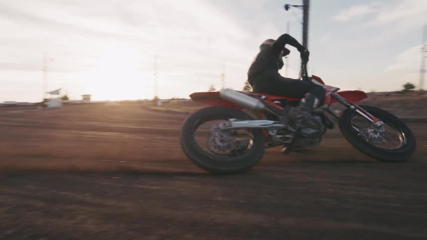 A dirt bike rider leans into a turn on a wide dirt track during sunset