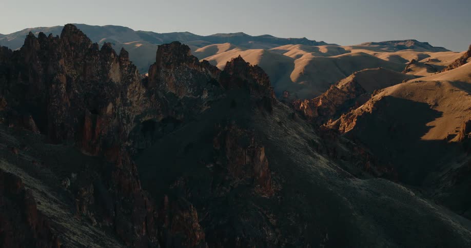 Dramatic dark jagged mountains and steep slopes are sharply contrasted by sunlit golden peaks and distant rolling hills under a clear blue sky highlighting the rugged terrain