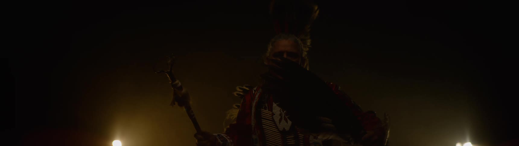 A native american person wearing a feathered headdress and traditional regalia dances in a dark hazy environment while holding a tomahawk and looking directly at the camera with a bright light source behind them