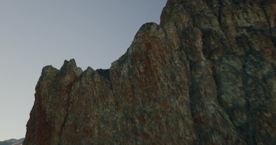 A rugged mountain with reddishbrown and dark gray rock faces rises against a gradient sky overlooking a shadowed valley with a dirt road and sunlit peaks