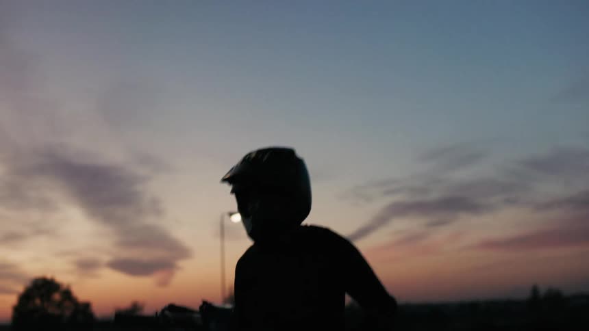 A silhouetted dirt bike rider in a helmet is positioned on a track with a vibrant sunset illuminating the sky behind them