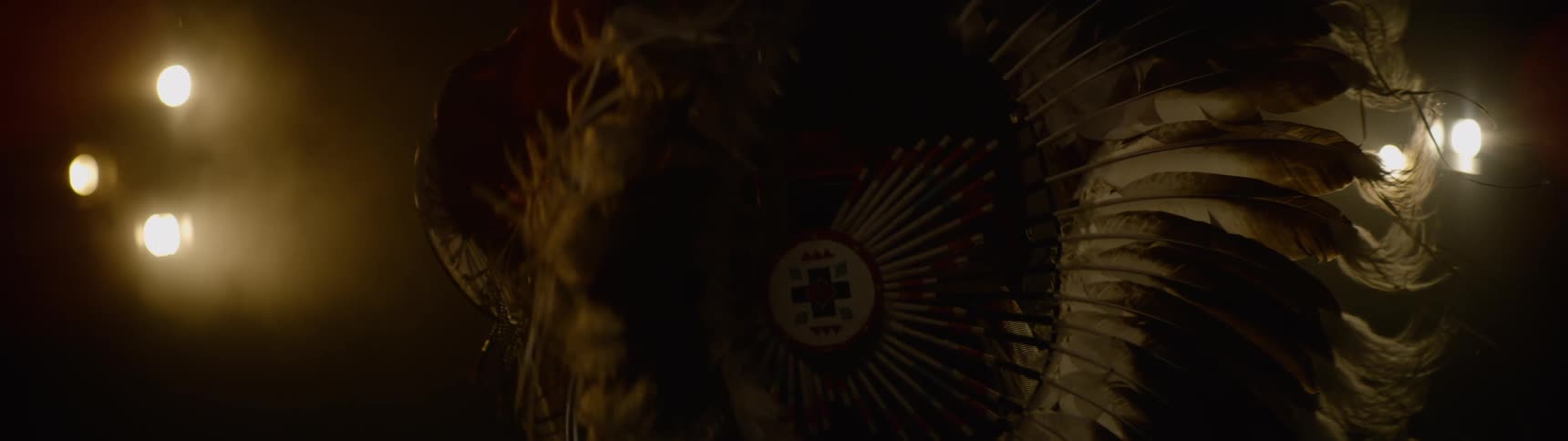 Native american dancer dances in ceremonial attire in a dim area lit by electric lights