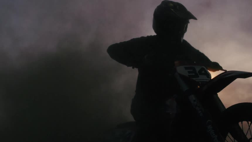 A motorcycle racer wearing a helmet warms up the bike releasing thick black smoke that fills the air at sunset