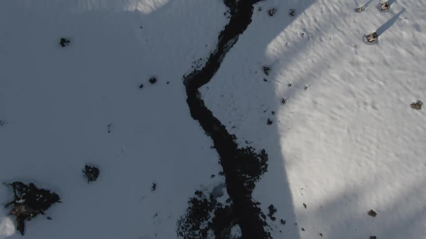 An aerial shot reveals a winding dark river flowing through a snowcovered landscape with scattered rocks and shadows