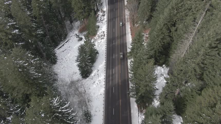 Aerial of vehicles moving on a long road in the middle of a woodland in daytime during winter