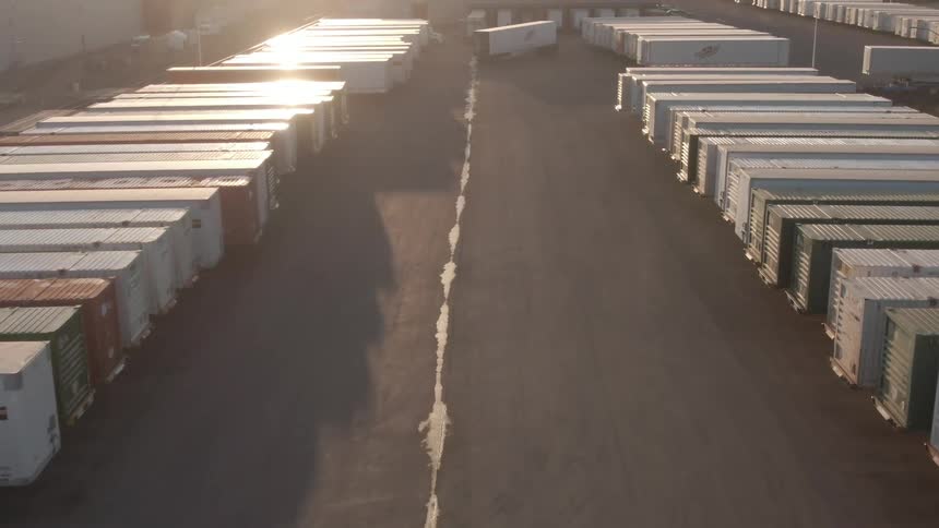 An aerial view shows a large industrial complex with numerous white shipping containers neatly parked in rows on a paved lot under a bright setting sun