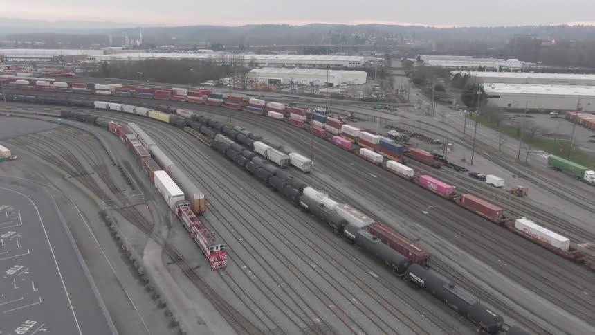 An aerial view flies over a rail yard lined with freight trains tanker cars and locomotives positioned beside a busy highway filled with moving vehicles