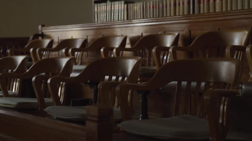Empty wooden jury seats line a courtroom beneath a shelf of law books