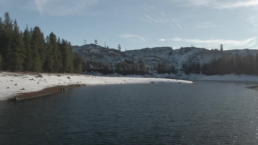 Snowcovered shoreline borders a calm lake surrounded by pine trees and distant hills
