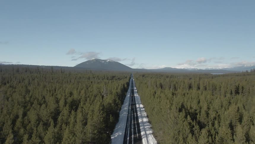 A snowcovered railway cuts through a vast pine forest toward distant mountains