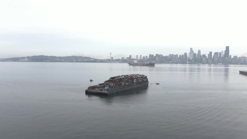 A barge in the ocean carrying a large load of scrap metals with a city skyline visible in the background at midday
