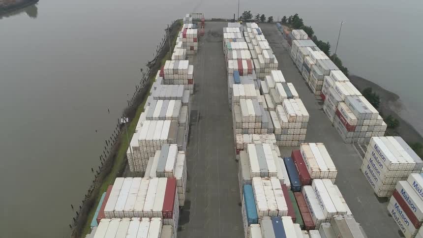 An aerial view captures a large port with numerous white shipping containers stacked in rows along a muddy river