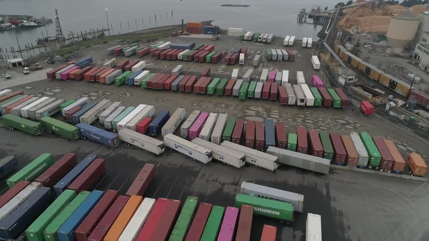 An aerial view captures a large shipping port filled with rows of multicolored cargo containers industrial buildings and a body of water with boats in the background