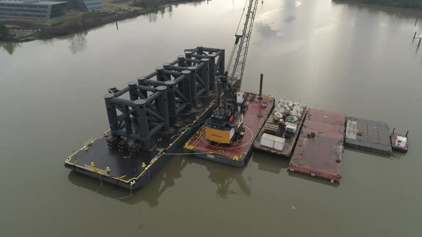A crane barge transports heavy industrial equipment along a calm river viewed from above