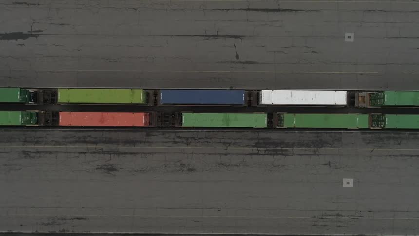An overhead shot shows a long train of colorful cargo containers on tracks positioned between two wide concrete strips