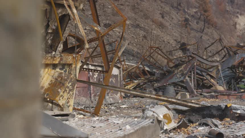 The charred and twisted remains of a building with rusted metal frames and debris lie in a desolate landscape against a backdrop of scorched hillsides and a hazy sky