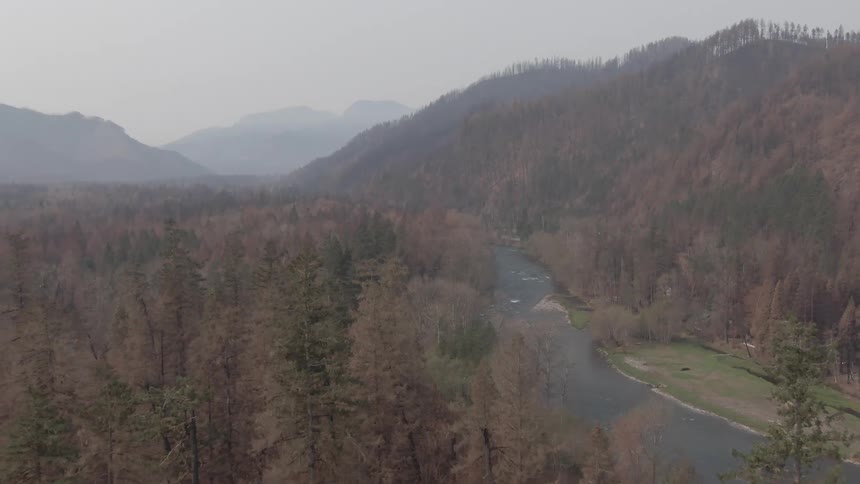 An aerial shot captures a winding river flowing through a wide valley surrounded by forested hills and mountains shrouded in haze with many of the trees appearing dry or dead