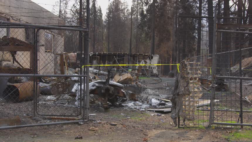 Burned structure and debris behind a damaged gate in a forested area