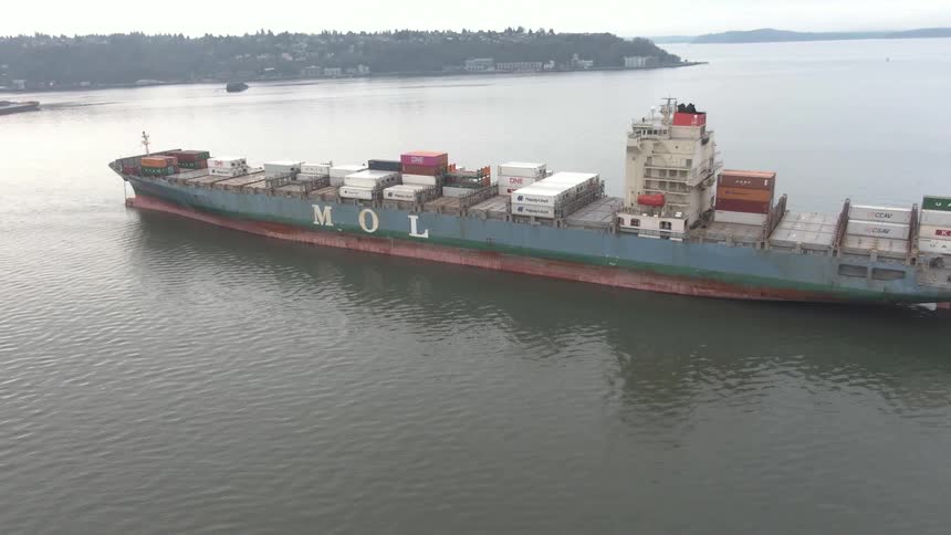 An aerial view captures a large mol container ship laden with cargo sailing across calm waters on an overcast day with a distant shoreline visible