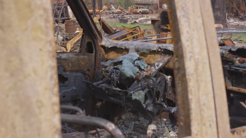 The rusted and charred interior of a burnedout vehicle is visible with debris and remnants of a destroyed structure in the background