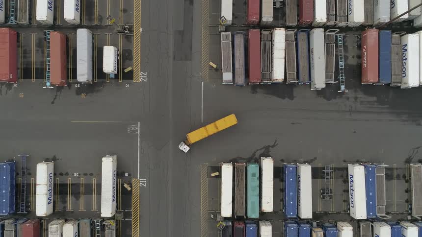 An aerial view shows a small container truck maneuvering through a cargo port yard as it transports a bright yellow shipping container past rows of parked freight units