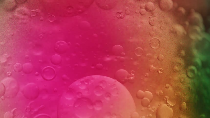 An extreme closeup abstract shot shows colorful pink orange and green oil bubbles of various sizes slowly floating and coalescing