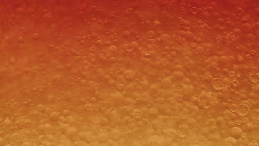 An extreme closeup shows many small bubbles suspended and slowly moving within a viscous orangecolored liquid creating an abstract textured surface