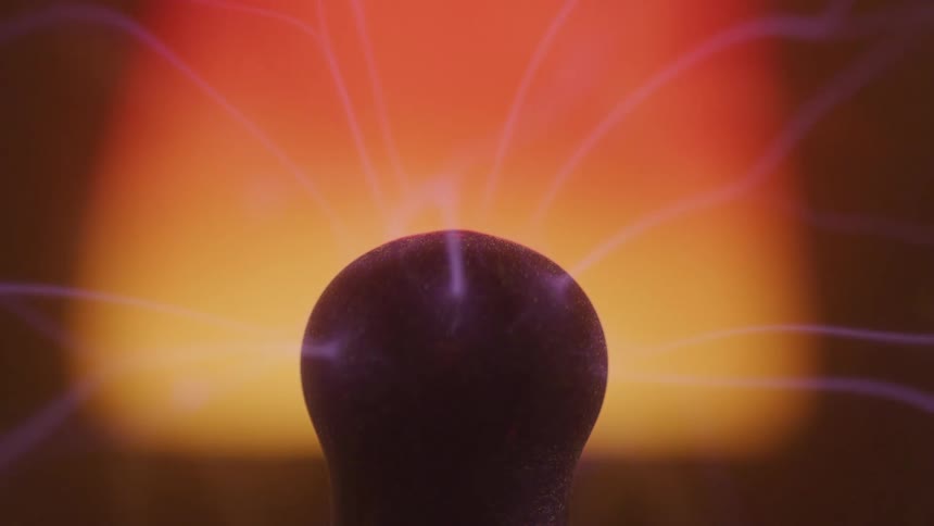 A closeup shot captures vibrant purple arcs of electricity moving around the central electrode of a plasma ball against a warm glowing orange background