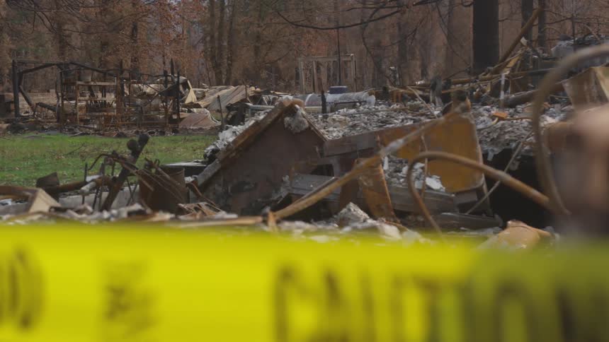 A foreground yellow caution tape is partially visible over a blurred wide shot of a firedamaged residential area with charred debris and skeletal structures