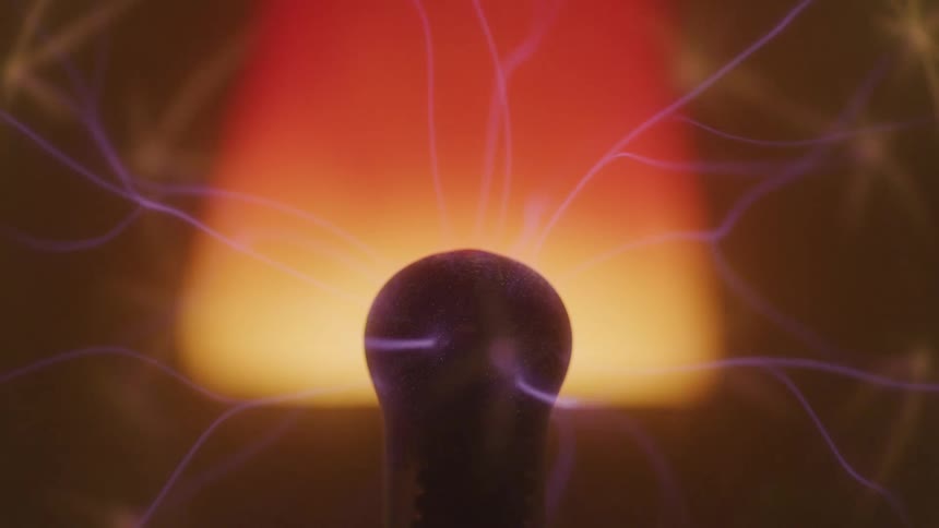 Purple electrical bolts radiate out away from aplasma ball that is silhouetted by a red and yellow ombre colored lamp in the background