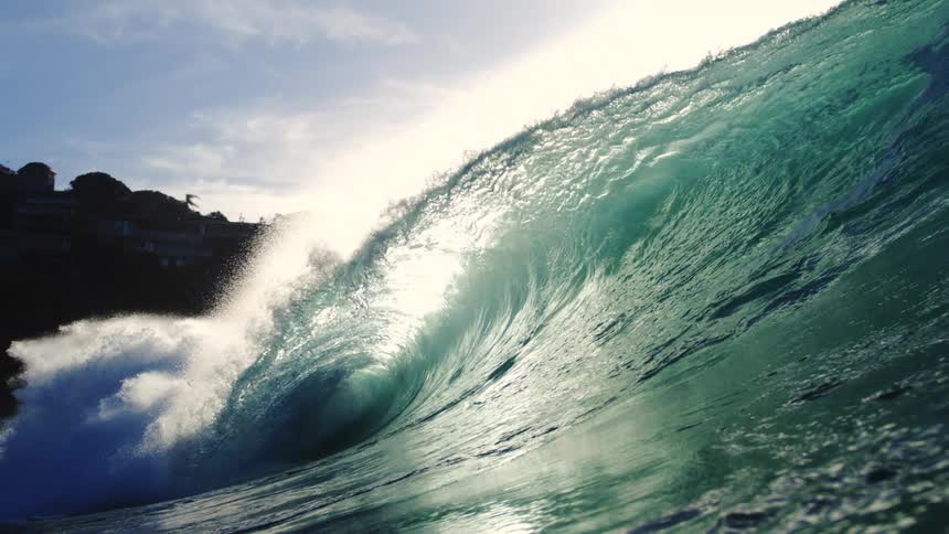 A large wave crests and breaks towards the right with bright sunlight reflecting off the water and buildings visible on a distant hillside