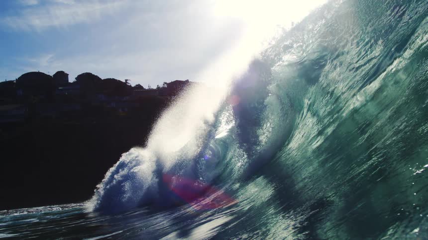 A powerful ocean wave crests with brilliant sun glare and lens flare revealing the transparent blue green water before submerging to show underwater bubbles and light filtering through the surface