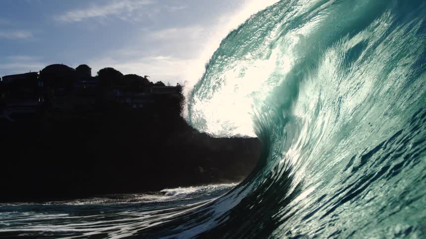 A powerful ocean wave with a curling crest illuminated by a bright sun and lens flares with a rocky cliff and houses visible in the distance
