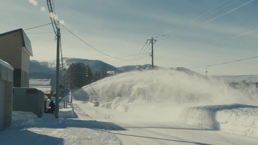 A small snowplow clears a snowcovered street in a village kicking up a large plume of snow towards the right side of the frame with mountains in the background under a bright sky
