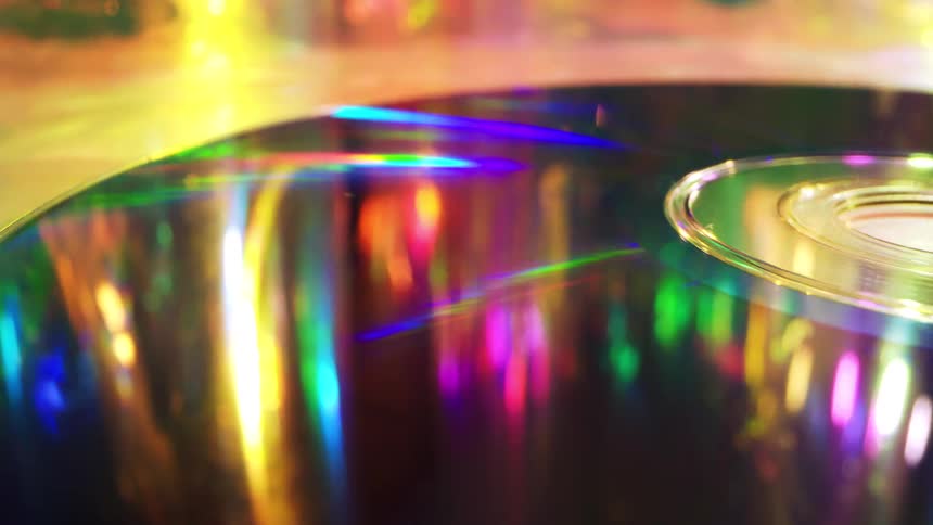 A compact disc lies on a dance floor reflecting streaks of party lights and golden streamers as dancers move around it in a shimmering club atmosphere