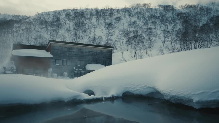 A steaming hot spring pool sits beside a wooden lodge surrounded by deep snow and forested mountains
