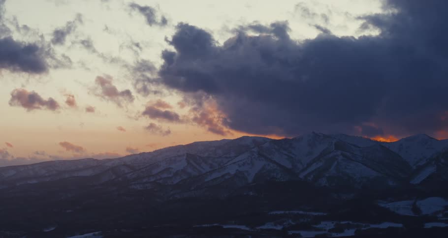 A vast snowcovered mountain range is dramatically illuminated by a sunset sky with a mix of dark and orangetinted clouds