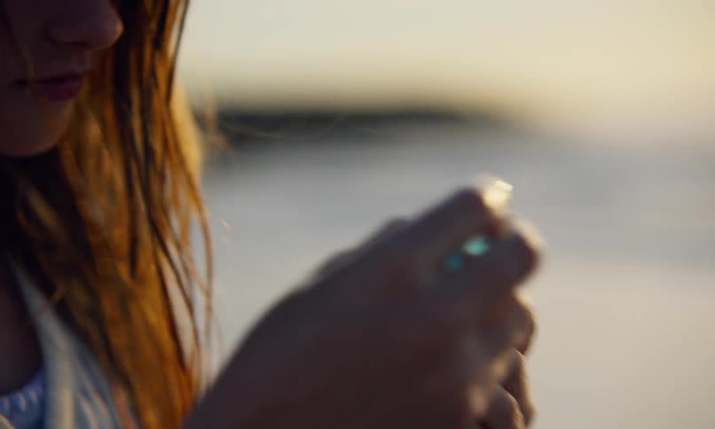 A young preteen girl with long, damp, red hair and a white dress is standing on the shoreline of a beach at sunset as she bends over to pick up a piece of translucent blue sea glass that just washed ashore.