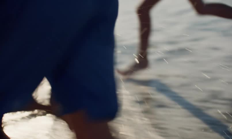 Focus on the legs of a group of children in swimsuits as they run together in the surf along the shore of a beach on a sunny day.