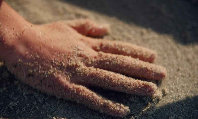 A close up of the texture and colors of grains of sand on a child's hand as they pat the ground while playing on a sunny day.
