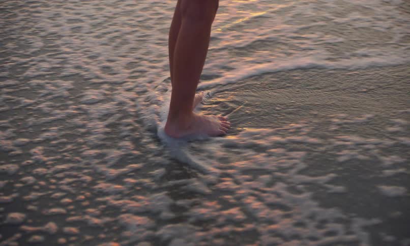 Close up of the lower legs and feet of a girl standing on the shoreline of a beach at sunset with her bare feet in the wet sand as the surf comes in and swirls around her feet leaving spots of sea foam behind.