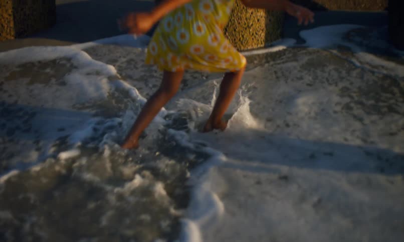 A young girl of color wearing a yellow flowered dress joyfully jumps in the surf along the shore near large coastal rocks and stone barriers.