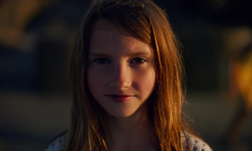 Portrait of a young Caucasian girl with long, copper-colored hair and a subtle smile standing on the beach at sunset looking directly at the camera as the camera pans back and a blurred figure walks behind her.