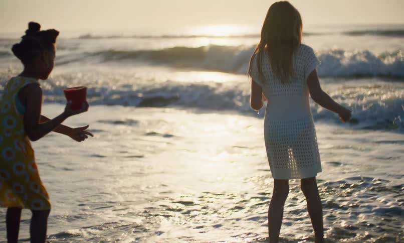 Slow motion of two young children playing and kicking the surf as it rolls in with the waves on a beach at sunset.