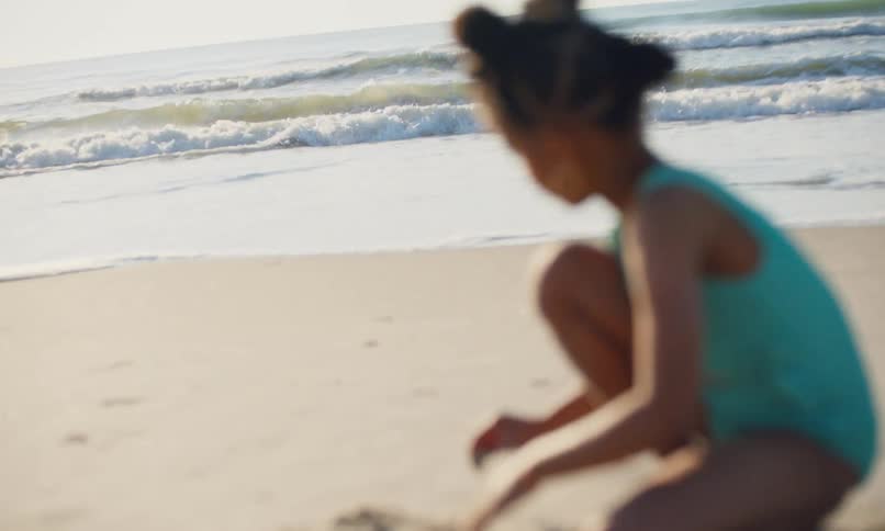 A little black girl in a blue swimsuit plays in the sand at the beach on a sunny day as ocean waves hit the shore.